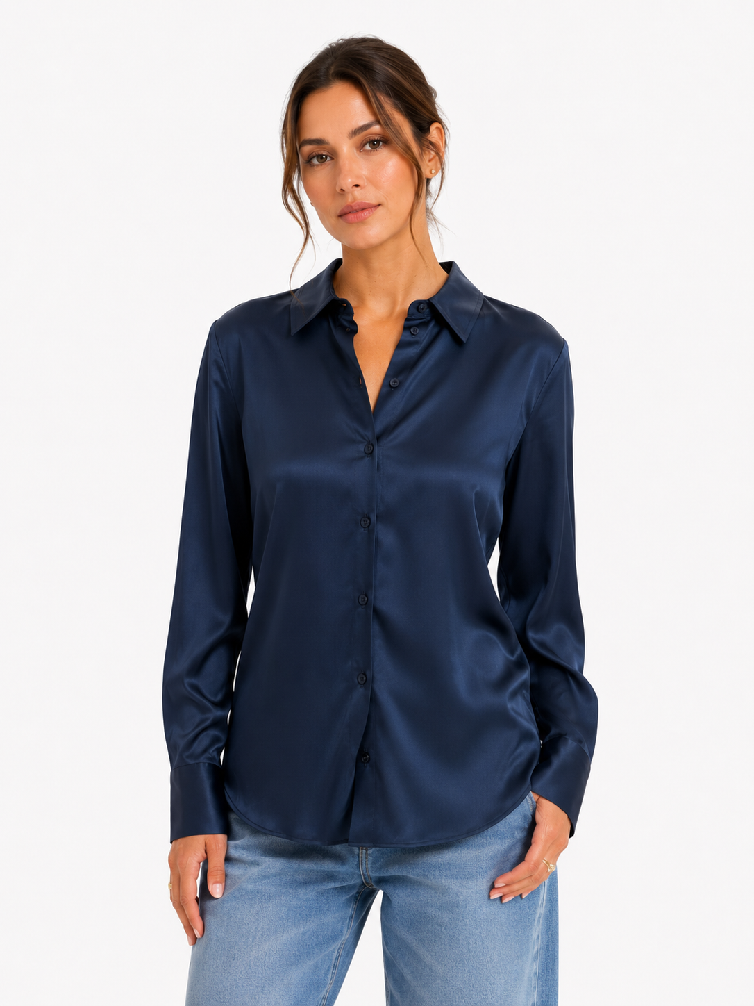 Eine Frau, die die Bluse „Kaia Dunkelblau“ aus fließendem Satin und hellblaue Jeans trägt, steht vor einem schlichten weißen Hintergrund und blickt mit ausdruckslosem Gesichtsausdruck in die Kamera.