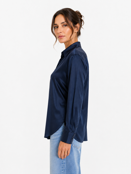 Eine Frau steht im Profil vor einem schlichten weißen Hintergrund und trägt die Bluse „Kaia Dunkelblau“ mit langen Ärmeln sowie hellblaue Jeans. Diese stilvolle Bluse wird mit Stolz in Portugal hergestellt.