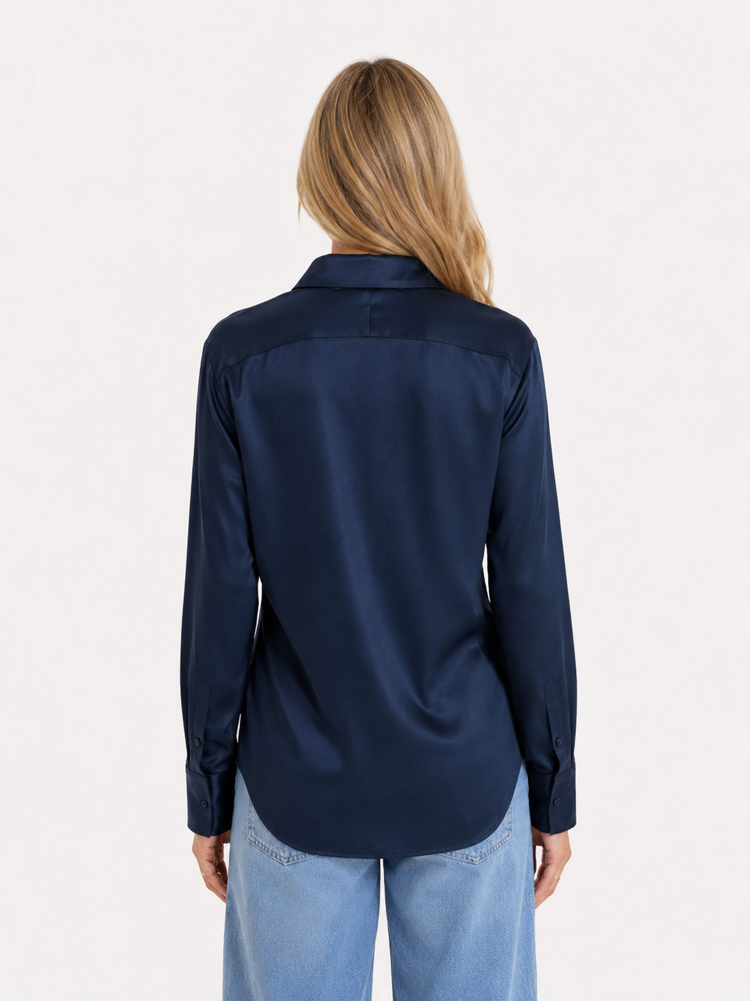 Eine Person mit langen blonden Haaren trägt die Bluse „Kaia Dunkelblau“ – ein fließend fallendes, marineblaues Satin-Langarmhemd aus portugiesischer Herstellung – sowie hellblaue Jeans und steht mit dem Rücken zur Kamera vor einem schlichten weißen Hintergrund.