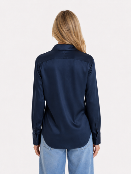 Eine Person mit langen blonden Haaren trägt die Bluse „Kaia Dunkelblau“ – ein fließend fallendes, marineblaues Satin-Langarmhemd aus portugiesischer Herstellung – sowie hellblaue Jeans und steht mit dem Rücken zur Kamera vor einem schlichten weißen Hintergrund.