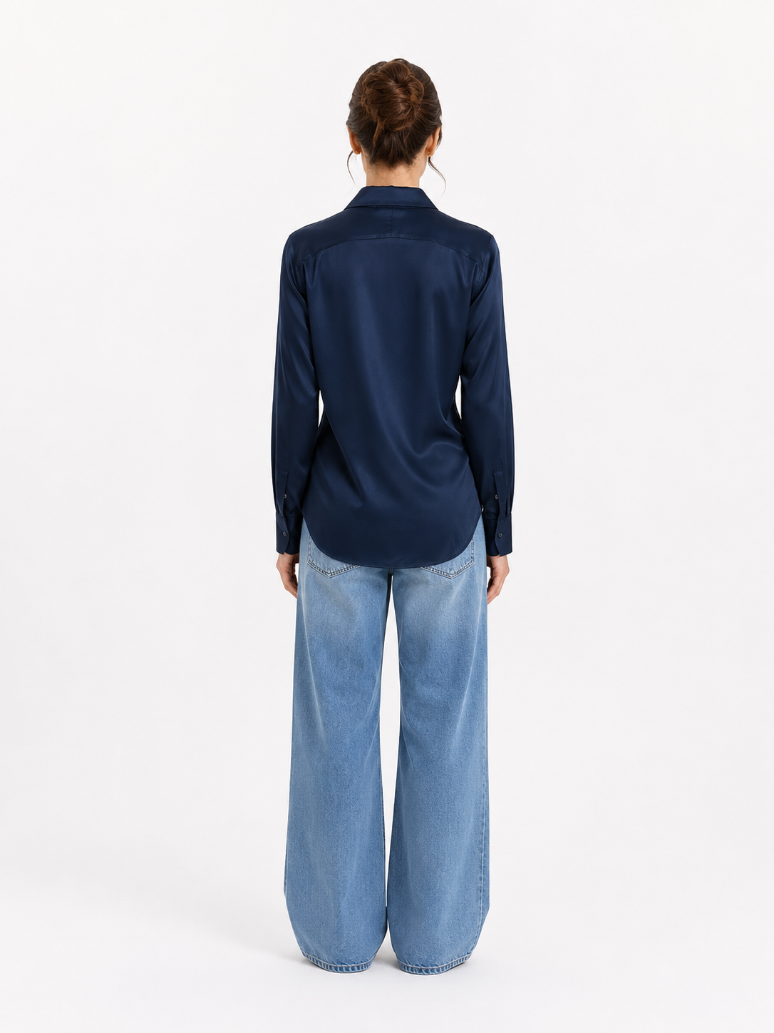Eine Frau steht mit dem Rücken zur Kamera und trägt die Bluse „Kaia Dunkelblau“ – eine langärmelige, dunkelblaue Satinbluse – sowie hellblaue Jeans mit weitem Bein. Ihr Haar ist hochgesteckt, und sie steht vor einem schlichten weißen Hintergrund.
