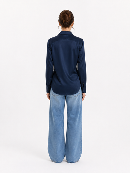 Eine Frau steht mit dem Rücken zur Kamera und trägt die Bluse „Kaia Dunkelblau“ – eine langärmelige, dunkelblaue Satinbluse – sowie hellblaue Jeans mit weitem Bein. Ihr Haar ist hochgesteckt, und sie steht vor einem schlichten weißen Hintergrund.