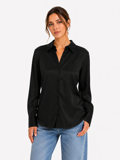 Eine Frau trägt die Bluse „Kaia Schwarz“ (hergestellt in Portugal), kombiniert mit einer hellblauen Jeans, und steht mit ausdruckslosem Gesicht vor einem schlichten weißen Hintergrund.