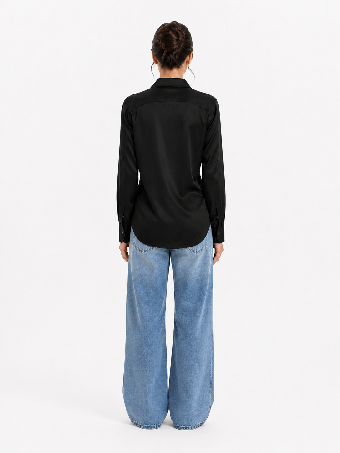 Eine Person steht mit dem Rücken zur Kamera und trägt die Bluse „Kaia Schwarz“ in Kombination mit einer blaufarbenen Jeans mit weitem Bein. Die Haare sind hochgesteckt, und die Person steht vor einem schlichten weißen Hintergrund. Der Look strahlt zurückhaltende Eleganz aus – jedes Detail ist sorgfältig gestaltet und stolz „Made in Portugal“.