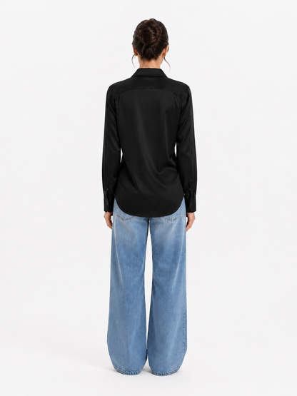 Eine Person steht mit dem Rücken zur Kamera und trägt die Bluse „Kaia Schwarz“ in Kombination mit einer blaufarbenen Jeans mit weitem Bein. Die Haare sind hochgesteckt, und die Person steht vor einem schlichten weißen Hintergrund. Der Look strahlt zurückhaltende Eleganz aus – jedes Detail ist sorgfältig gestaltet und stolz „Made in Portugal“.