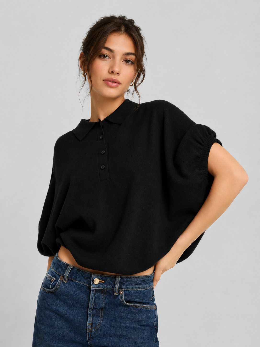 Eine Frau steht mit einem Arm in die Hüfte gestützt, trägt ein lockeres schwarzes Polohemd mit Kragen von „Cowboy Polo Knit Black“ und eine blaue Jeans und blickt vor einem schlichten hellgrauen Hintergrund selbstbewusst in die Kamera.