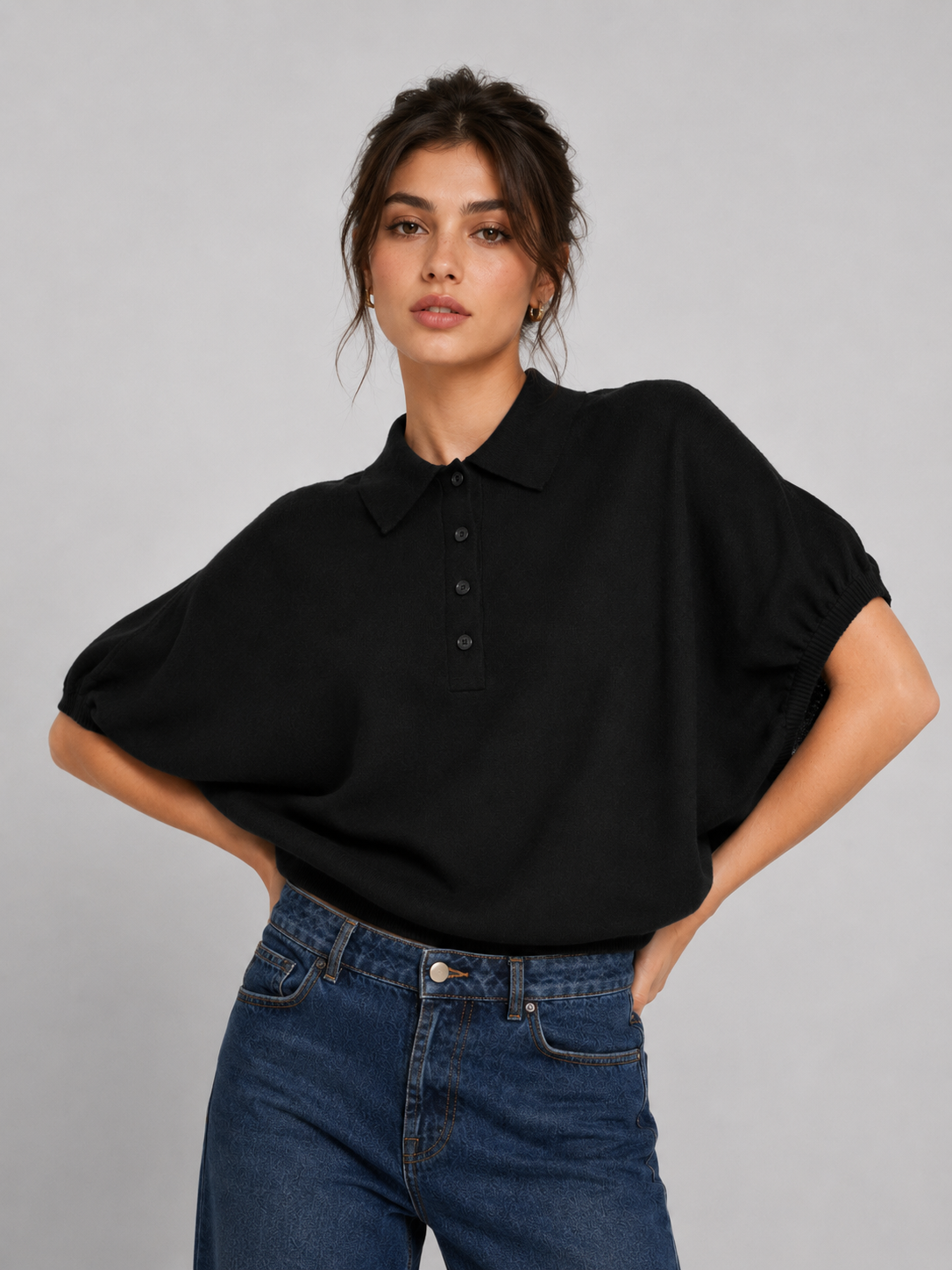 Eine Frau, die ein locker sitzendes schwarzes „Cowboy Polo Knit“-Shirt und eine blaue Jeans trägt, steht mit den Händen in den Hüften vor einem schlichten hellgrauen Hintergrund.