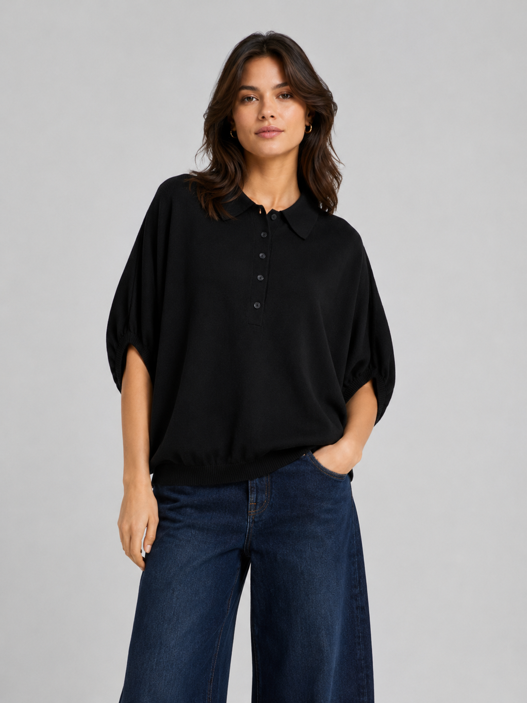 Eine Frau steht vor einem schlichten hellgrauen Hintergrund, blickt in die Kamera, hat eine Hand in der Tasche und trägt ein lockeres schwarzes Hemd mit Kragen und Knöpfen, das an das Modell „Cowboy Polo Knit Black“ erinnert, sowie eine weite dunkelblaue Jeans.
