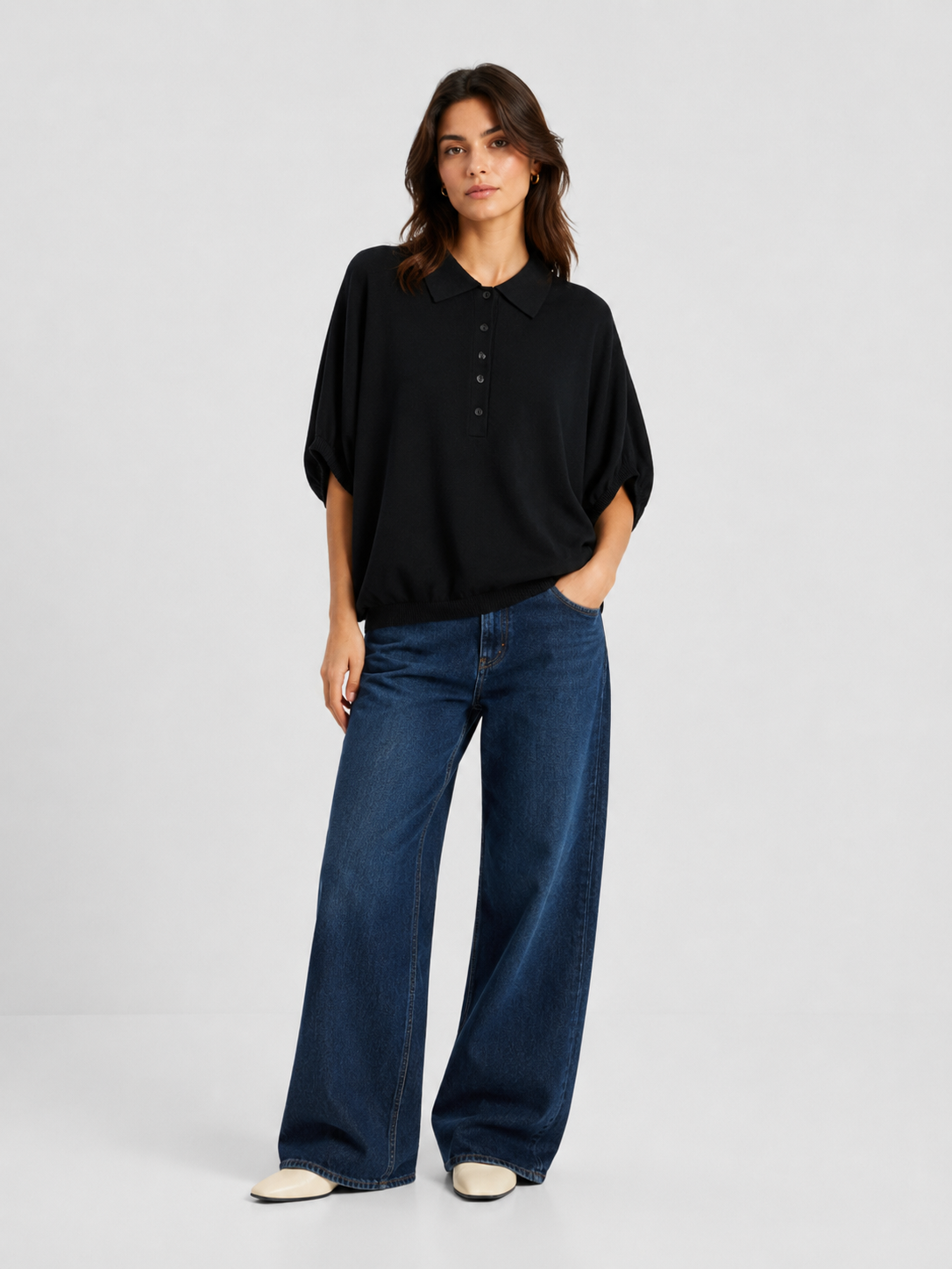 Eine Person steht vor einem schlichten Hintergrund und trägt ein locker sitzendes schwarzes Cowboy-Polo-Shirt aus Strick, eine blaue Jeans mit weitem Bein und weiße Schuhe; eine Hand steckt in der Hosentasche, ihr Gesichtsausdruck ist neutral.