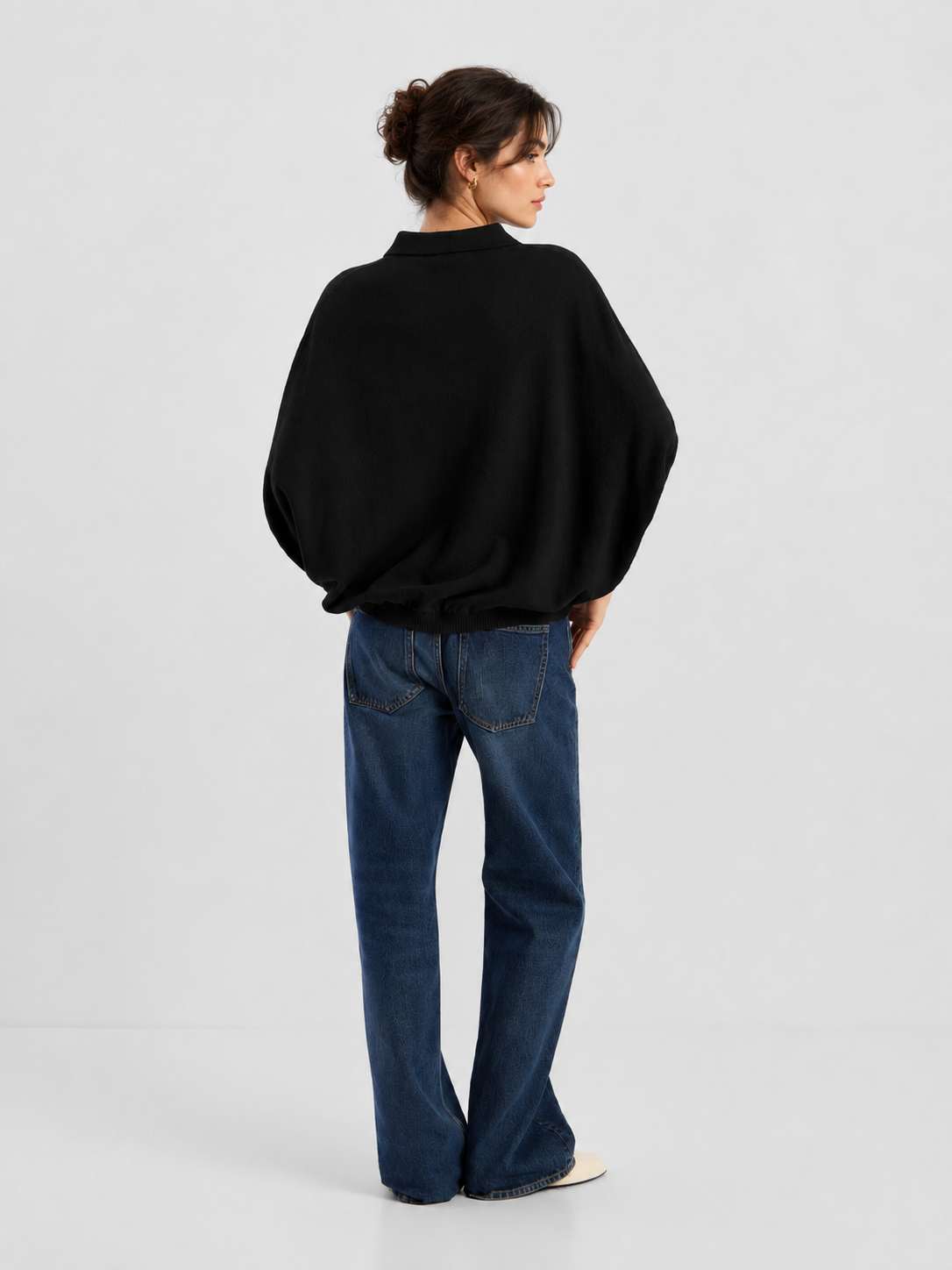 Eine Frau steht mit dem Rücken zur Kamera und trägt ein lockeres schwarzes „Cowboy Polo Knit“-Shirt mit Fledermausärmeln, blaue Jeans und weiße Schuhe. Ihr Haar ist hochgesteckt, und sie steht vor einem schlichten, hellen Hintergrund.