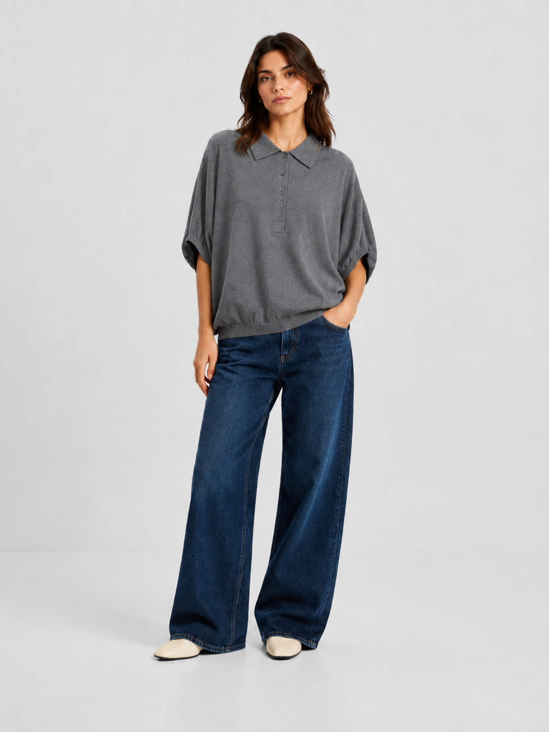 Eine Person steht vor einem schlichten Hintergrund und trägt ein locker sitzendes graues „Cowboy Polo Knit“-Shirt, eine blaue Jeans mit weitem Bein und weiße Schuhe; eine Hand steckt in der Hosentasche.