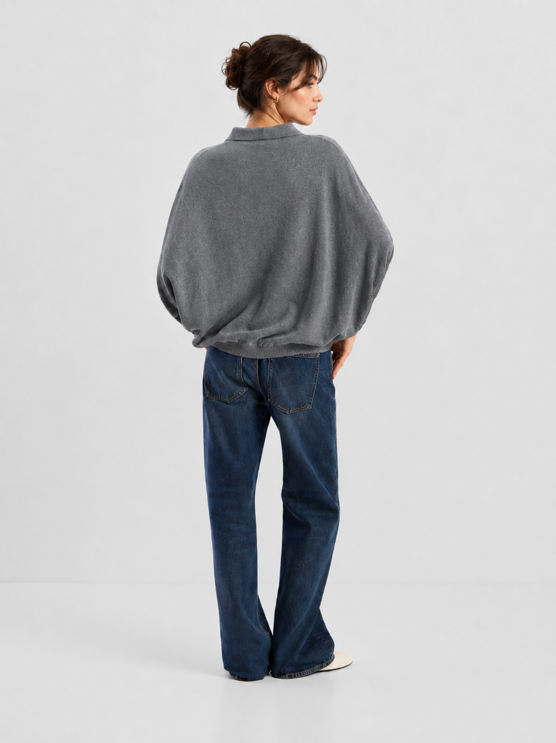 Eine Person steht mit dem Rücken zur Kamera und trägt das „Cowboy Polo Knit Grey“ zusammen mit blauen Jeans und weißen Schuhen. Die Haare sind hochgesteckt, und die Person steht vor einem schlichten, hellen Hintergrund.