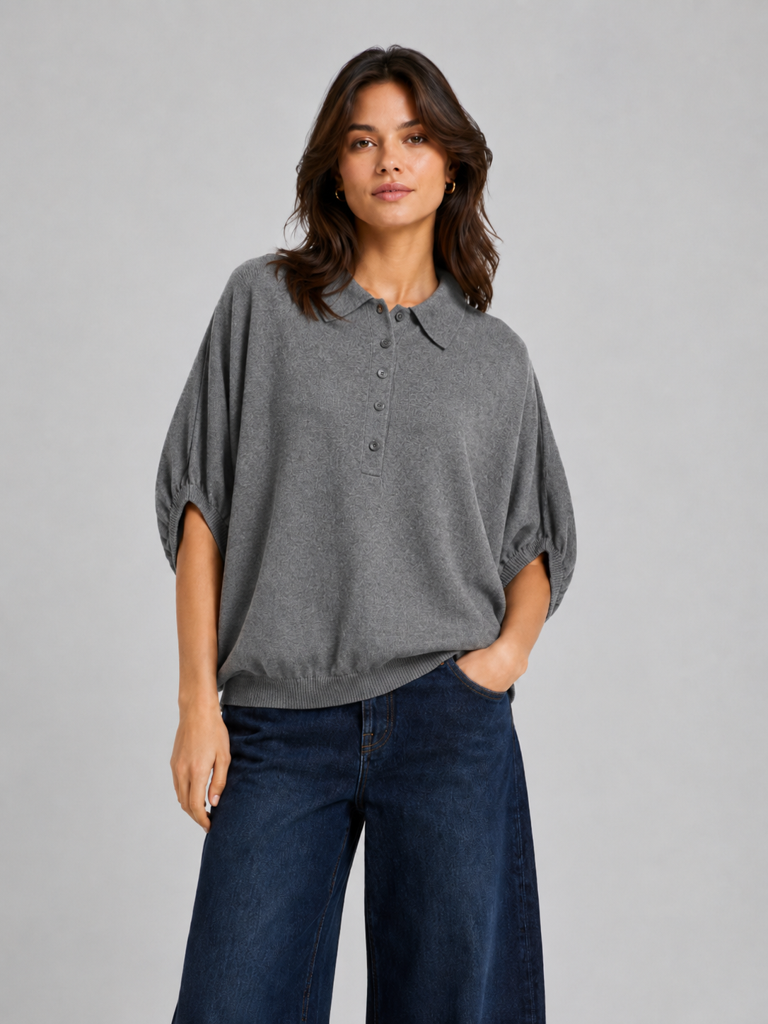 Vor einem schlichten Hintergrund steht eine Frau, die das edle „Cowboy Polo Knit Grey“ trägt – ein locker sitzendes, graues Kurzarm-Poloshirt, das an einen Pullover aus Merinowolle und Kaschmir erinnert – dazu eine dunkelblaue Jeans mit weitem Bein. Sie hat welliges braunes Haar und einen neutralen Gesichtsausdruck; eine Hand steckt in ihrer Hosentasche.