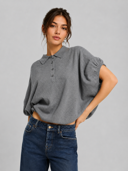 Eine Frau in einem locker sitzenden, grauen Cowboy-Polo-Strickhemd mit Kragen und blauen Jeans steht mit einer Hand in der Hüfte und posiert vor einem schlichten hellgrauen Hintergrund.