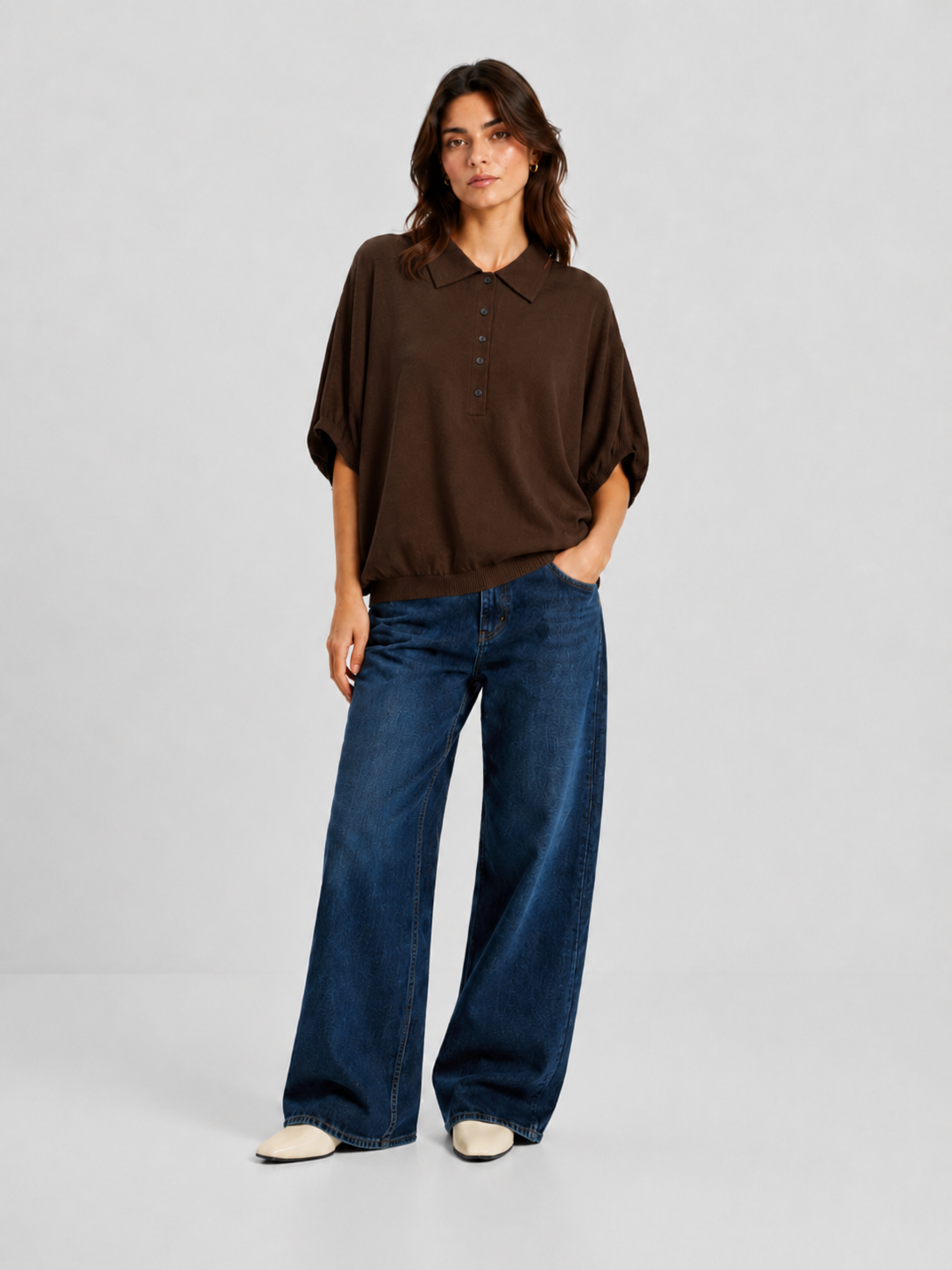 Vor einem schlichten Hintergrund steht eine Frau, die ein locker sitzendes, braunes „Cowboy Polo Knit“-Oberteil aus luxuriöser Merinowolle und Kaschmir, eine blaue Jeans mit weitem Bein und weiße Schuhe trägt. Sie hat schulterlanges braunes Haar und nimmt eine entspannte Haltung ein, wobei sie eine Hand in die Tasche gesteckt hat.