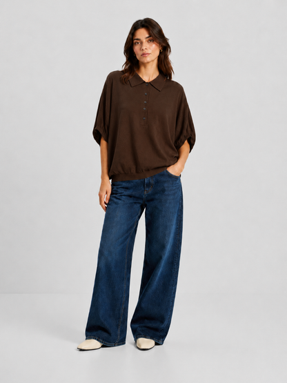 Vor einem schlichten Hintergrund steht eine Frau, die ein locker sitzendes, braunes „Cowboy Polo Knit“-Oberteil aus luxuriöser Merinowolle und Kaschmir, eine blaue Jeans mit weitem Bein und weiße Schuhe trägt. Sie hat schulterlanges braunes Haar und nimmt eine entspannte Haltung ein, wobei sie eine Hand in die Tasche gesteckt hat.