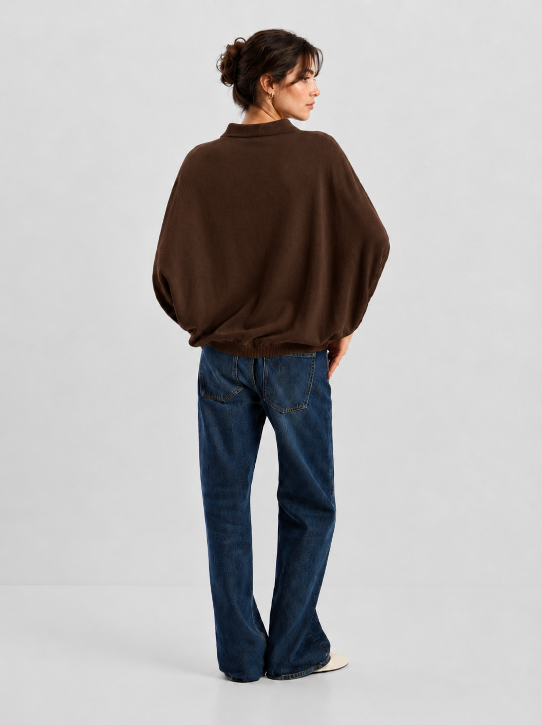 Eine Person steht mit dem Rücken zur Kamera und trägt ein locker sitzendes, braunes „Cowboy Polo Knit“-Shirt aus einer luxuriösen Merino-Kaschmir-Mischung, dazu blaue Jeans und weiße Schuhe vor einem schlichten, hellen Hintergrund. Die Haare sind hochgesteckt.