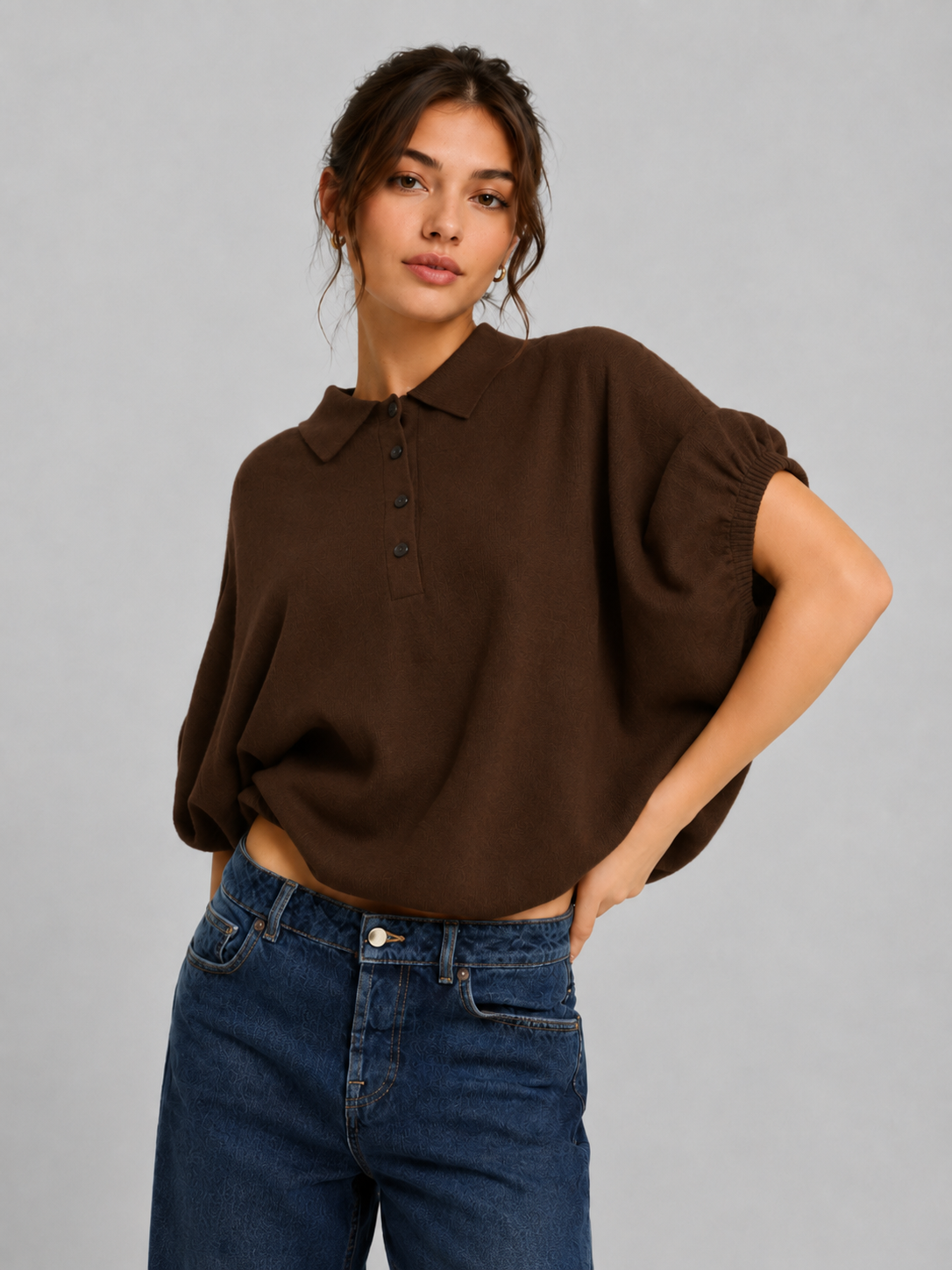 Eine Frau mit braunem, welligem Haar trägt ein locker sitzendes, kurzärmeliges „Cowboy Polo Knit Brown“-Hemd aus Merinowolle und Kaschmir, kombiniert mit einer blauen Jeans. Sie steht mit einer Hand in der Hüfte vor einem schlichten grauen Hintergrund.