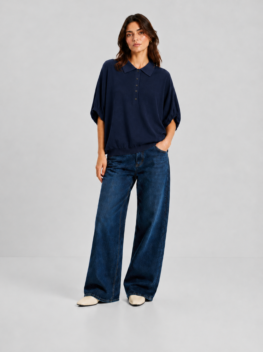 Eine Person steht vor einem hellgrauen Hintergrund und trägt ein locker sitzendes „Cowboy Polo Knit Navy“ – ein marineblaues Hemd mit Kragen aus luxuriösem Feinstrick –, dazu eine blaue Jeans mit weitem Bein und weiße Schuhe; eine Hand steckt lässig in der Tasche.