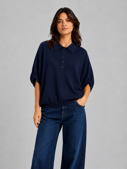 Eine Person, die ein locker sitzendes, marineblaues „Cowboy Polo Knit“-Hemd mit Kragen und Knöpfen sowie eine blaue Jeans mit weitem Bein trägt, steht vor einem schlichten grauen Hintergrund und blickt mit einer Hand in der Hosentasche in die Kamera.