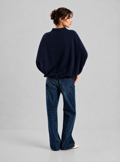Eine Frau steht mit dem Rücken zur Kamera und trägt ein „Cowboy Polo Knit Navy“ – luxuriös und elegant – dazu blaue Jeans und weiße Schuhe; ihr Haar ist ordentlich hochgesteckt, vor einem schlichten hellgrauen Hintergrund.