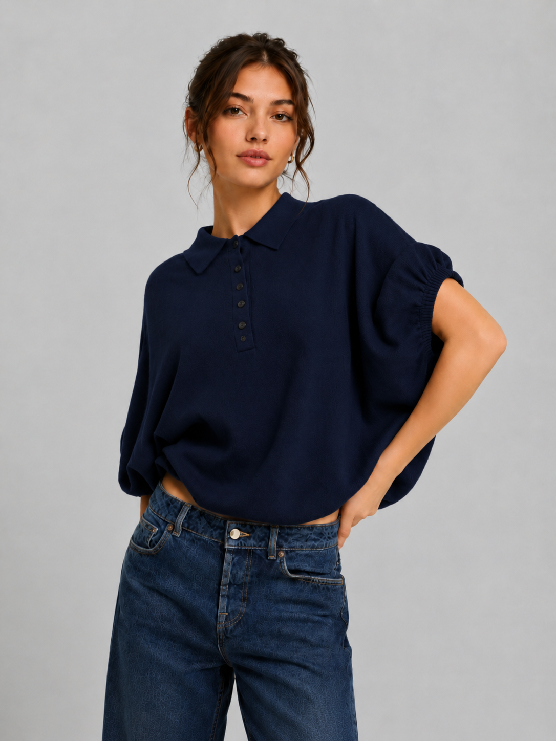 Eine Frau, die ein locker sitzendes „Cowboy Polo Knit Navy“ und eine blaue Jeans trägt, steht mit einer Hand in der Hüfte vor einem schlichten hellgrauen Hintergrund.