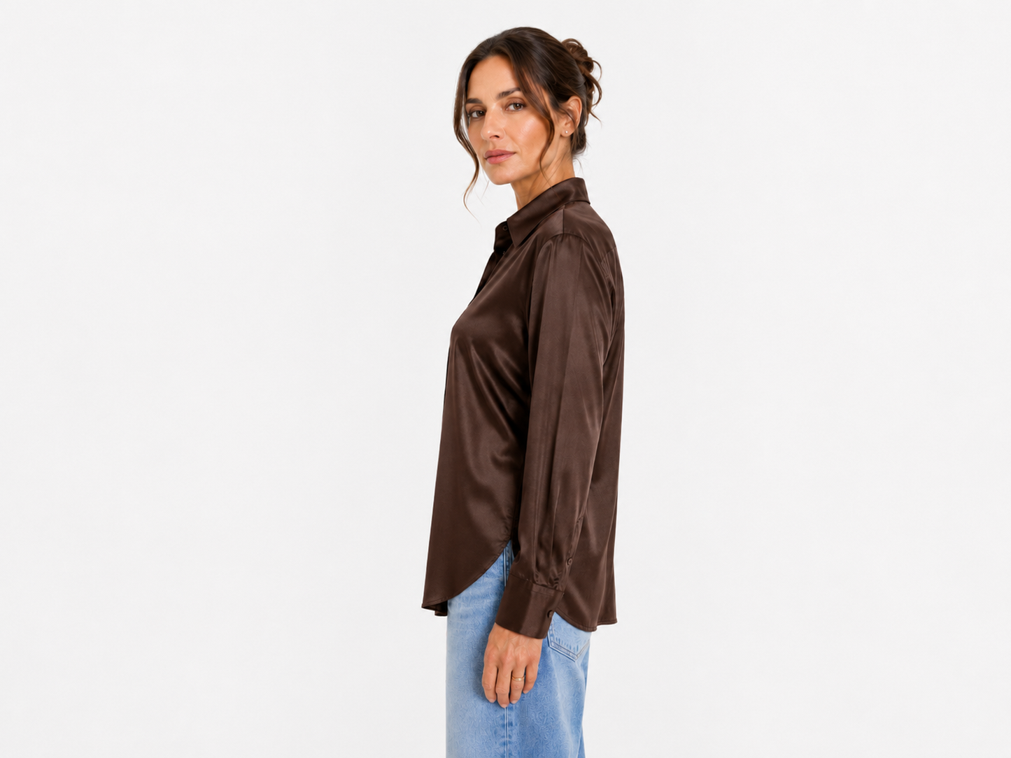 Eine Frau steht im Profil vor einem weißen Hintergrund. Sie trägt die langärmelige Bluse „Kaia Braun“ und hellblaue Jeans, ihr Haar ist zu einer lockeren Hochsteckfrisur gestylt. Hergestellt in Portugal.
