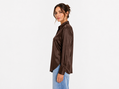 Eine Frau steht im Profil vor einem weißen Hintergrund. Sie trägt die langärmelige Bluse „Kaia Braun“ und hellblaue Jeans, ihr Haar ist zu einer lockeren Hochsteckfrisur gestylt. Hergestellt in Portugal.