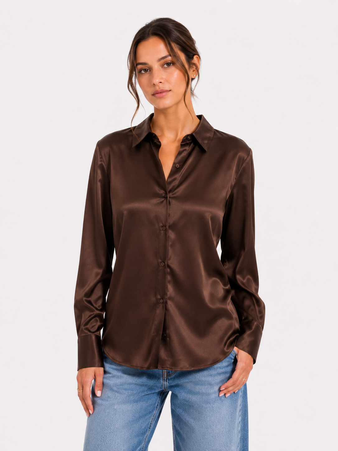 Eine Frau, die die Bluse „Kaia Braun“ – eine glänzende braune Satinbluse – und eine blaue Jeans trägt, steht mit ausdruckslosem Gesicht vor einem schlichten weißen Hintergrund. Diese elegante Bluse wird in Portugal hergestellt.