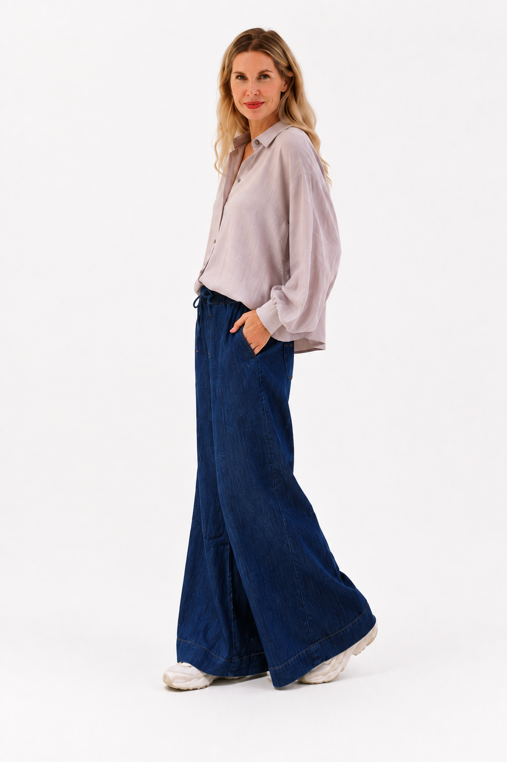 Eine Frau steht im Profil und trägt eine lockere, helle Bluse, eine Hose „Anna“ in Dunkelblau im Denim-Look mit weitem Bein sowie weiße Schuhe. Sie hat langes, welliges blondes Haar und blickt mit einem leichten Lächeln in die Kamera, während sie die Hände in den Taschen hat.