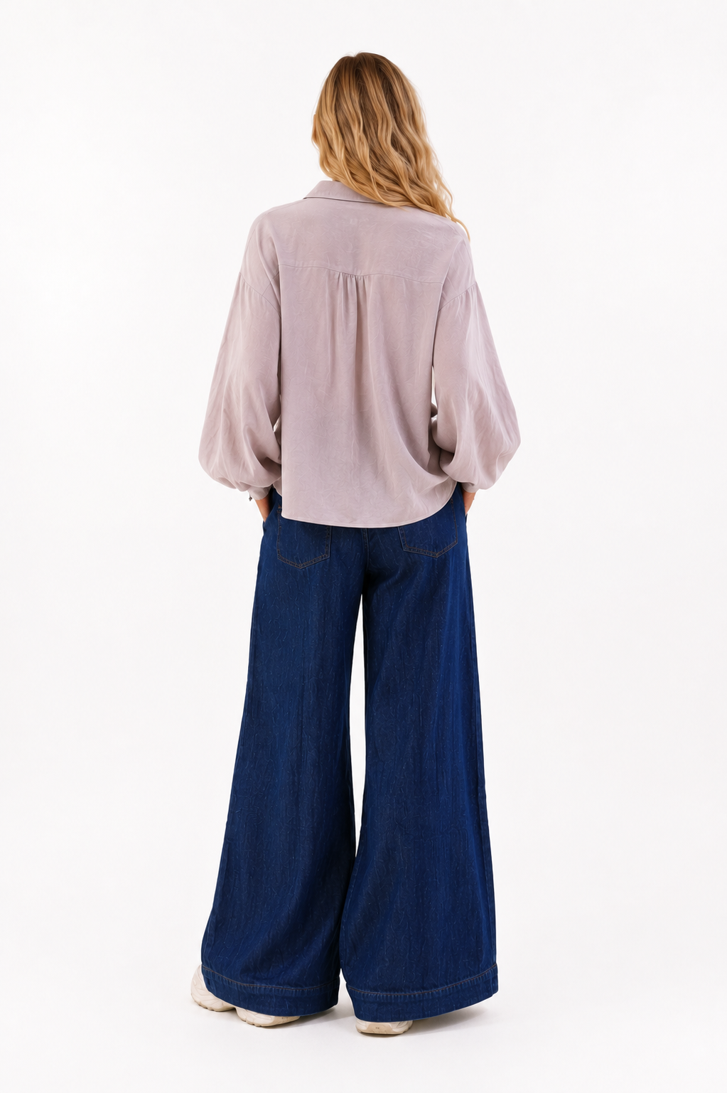 Eine Person steht mit dem Rücken zur Kamera und trägt ein lockeres, helles Langarmhemd sowie die Hose „Anna Dark Blue“ im klassischen Denim-Stil mit weitem Bein. Ihr blondes, welliges Haar fällt ihr über die Schultern. Der Hintergrund ist schlicht und weiß.