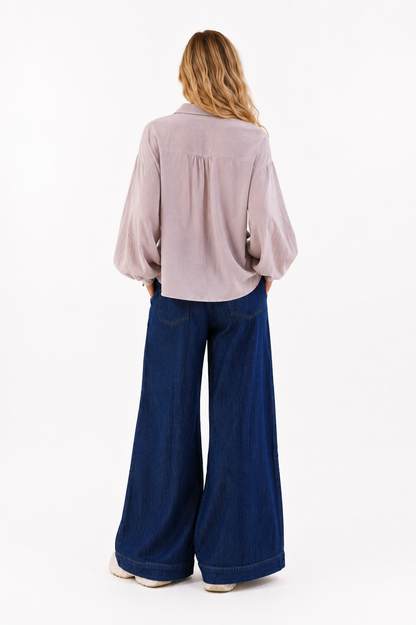 Eine Person steht mit dem Rücken zur Kamera und trägt ein lockeres, helles Langarmhemd sowie die Hose „Anna Dark Blue“ im klassischen Denim-Stil mit weitem Bein. Ihr blondes, welliges Haar fällt ihr über die Schultern. Der Hintergrund ist schlicht und weiß.