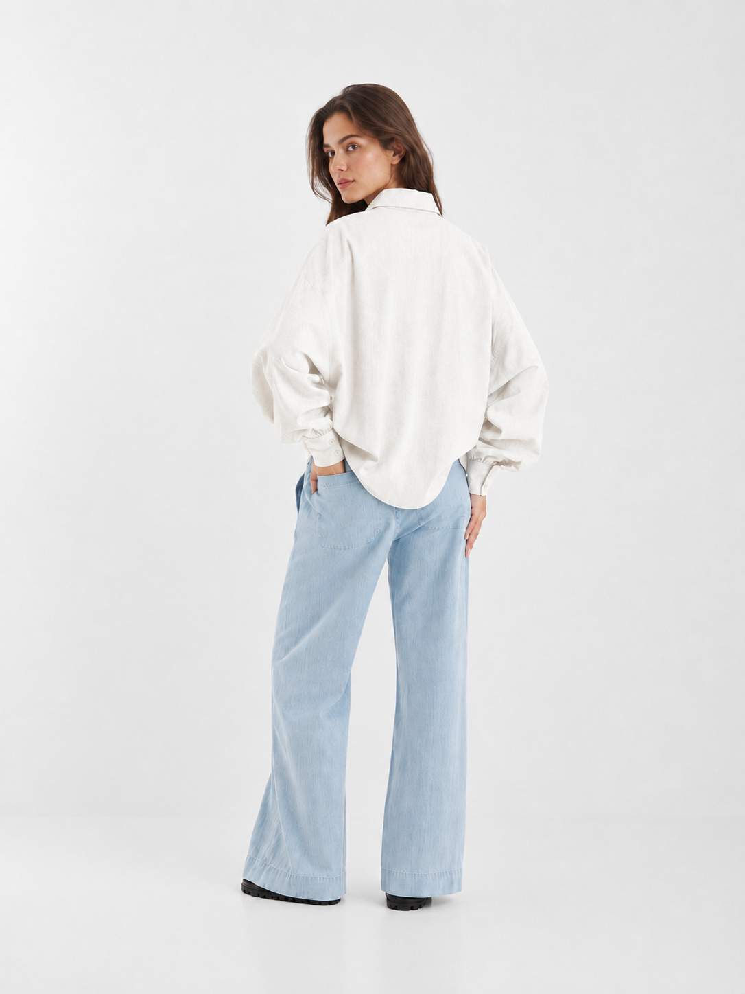 Eine Frau steht mit dem Rücken zur Kamera und blickt über die Schulter. Sie trägt eine locker sitzende Bluse „Antonia Slim Flannel Offwhite“ und eine hellblaue Jeans mit weitem Bein vor einem schlichten weißen Hintergrund.
