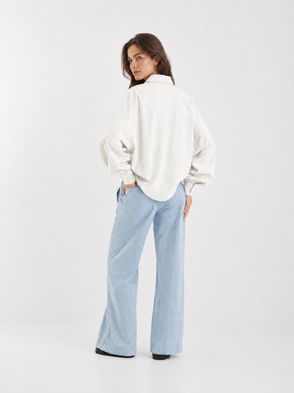 Eine Frau steht mit dem Rücken zur Kamera und blickt über die Schulter. Sie trägt eine locker sitzende Bluse „Antonia Slim Flannel Offwhite“ und eine hellblaue Jeans mit weitem Bein vor einem schlichten weißen Hintergrund.