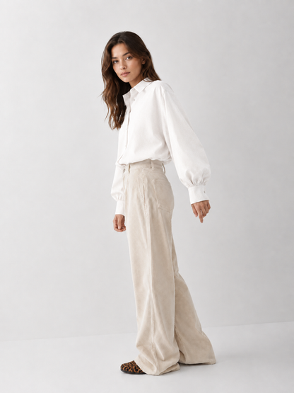 Eine Frau, die eine locker sitzende weiße Bluse „Antonia Slim Flannel Offwhite“ und eine beige Cordhose mit weitem Bein trägt, steht seitlich vor einem schlichten hellen Hintergrund und blickt in die Kamera. Diese Damenbluse aus Premium-Flanell wird in Portugal hergestellt und bietet sowohl Tragekomfort als auch zeitlosen Stil.