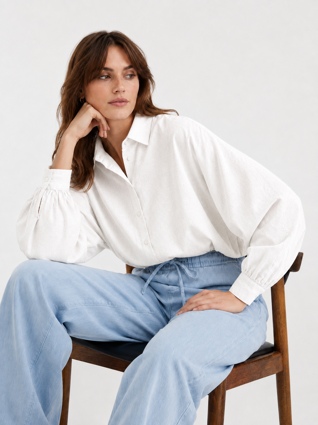 Eine Frau sitzt auf einem Holzstuhl und trägt die Bluse „Antonia Slim Flannel Offwhite“ mit weiten Ärmeln sowie eine hellblaue Hose mit weitem Bein. Vor einem schlichten Hintergrund blickt sie mit entspanntem Gesichtsausdruck zur Seite. Diese Premium-Flanell-Damenbluse wird in Portugal meisterhaft gefertigt und vereint Komfort mit zeitlosem Stil.