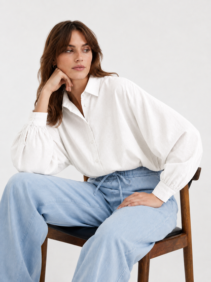 Eine Frau sitzt auf einem Holzstuhl und trägt die Bluse „Antonia Slim Flannel Offwhite“ mit weiten Ärmeln sowie eine hellblaue Hose mit weitem Bein. Vor einem schlichten Hintergrund blickt sie mit entspanntem Gesichtsausdruck zur Seite. Diese Premium-Flanell-Damenbluse wird in Portugal meisterhaft gefertigt und vereint Komfort mit zeitlosem Stil.