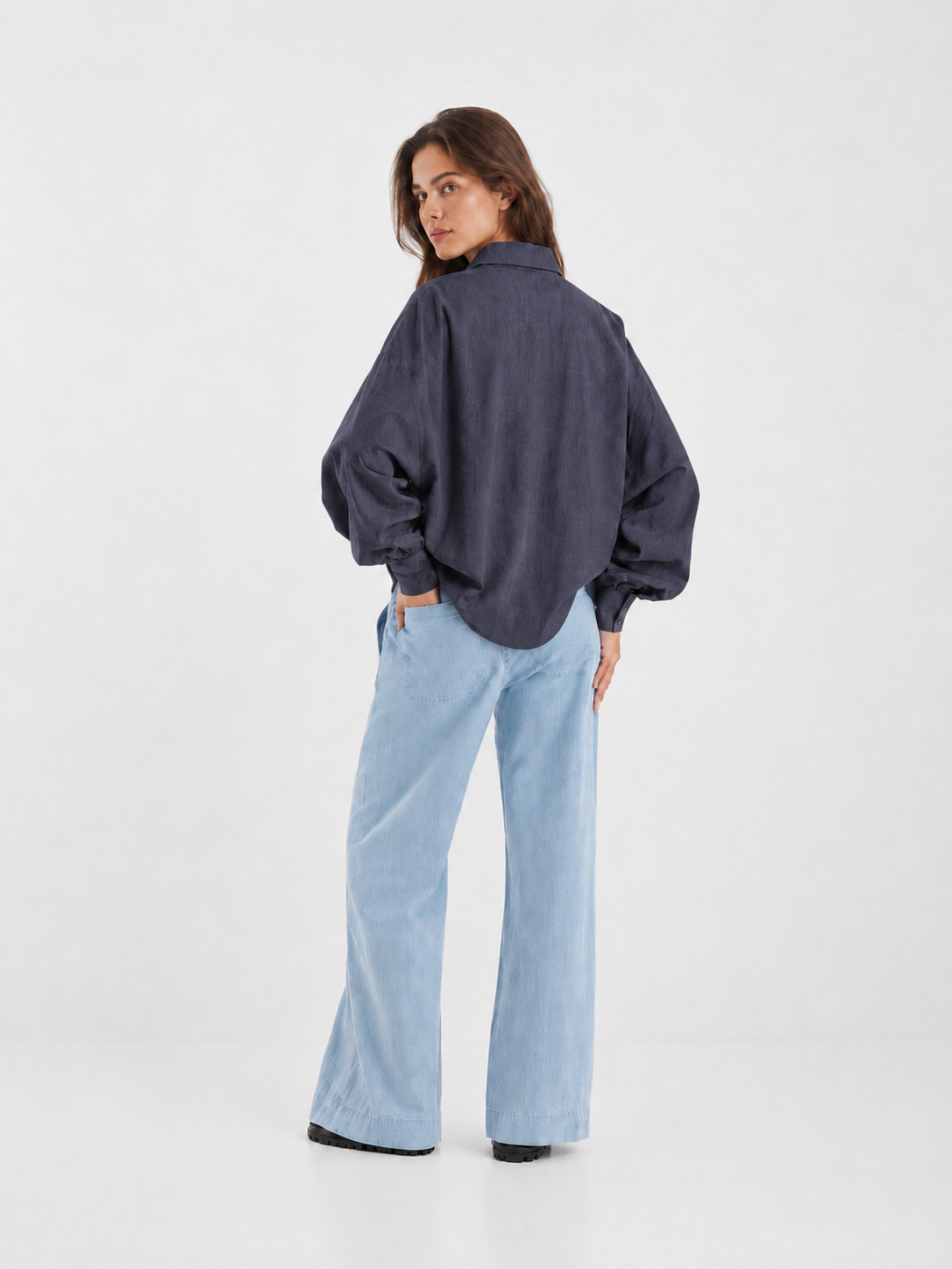 Eine Frau steht mit dem Rücken zur Kamera und blickt über ihre Schulter. Sie trägt eine locker sitzende, dunkelanthrazitfarbene Bluse „Antonia Slim Flannel“ und eine hellblaue Hose mit weitem Bein. Der Hintergrund ist schlicht und weiß.