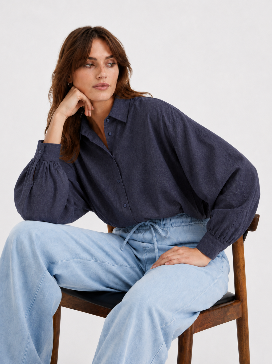 Eine Person sitzt auf einem Holzstuhl, trägt die Bluse „Antonia Slim Flannel Anthra“ mit Puffärmeln und eine hellblaue Hose mit weitem Bein und schaut zur Seite.