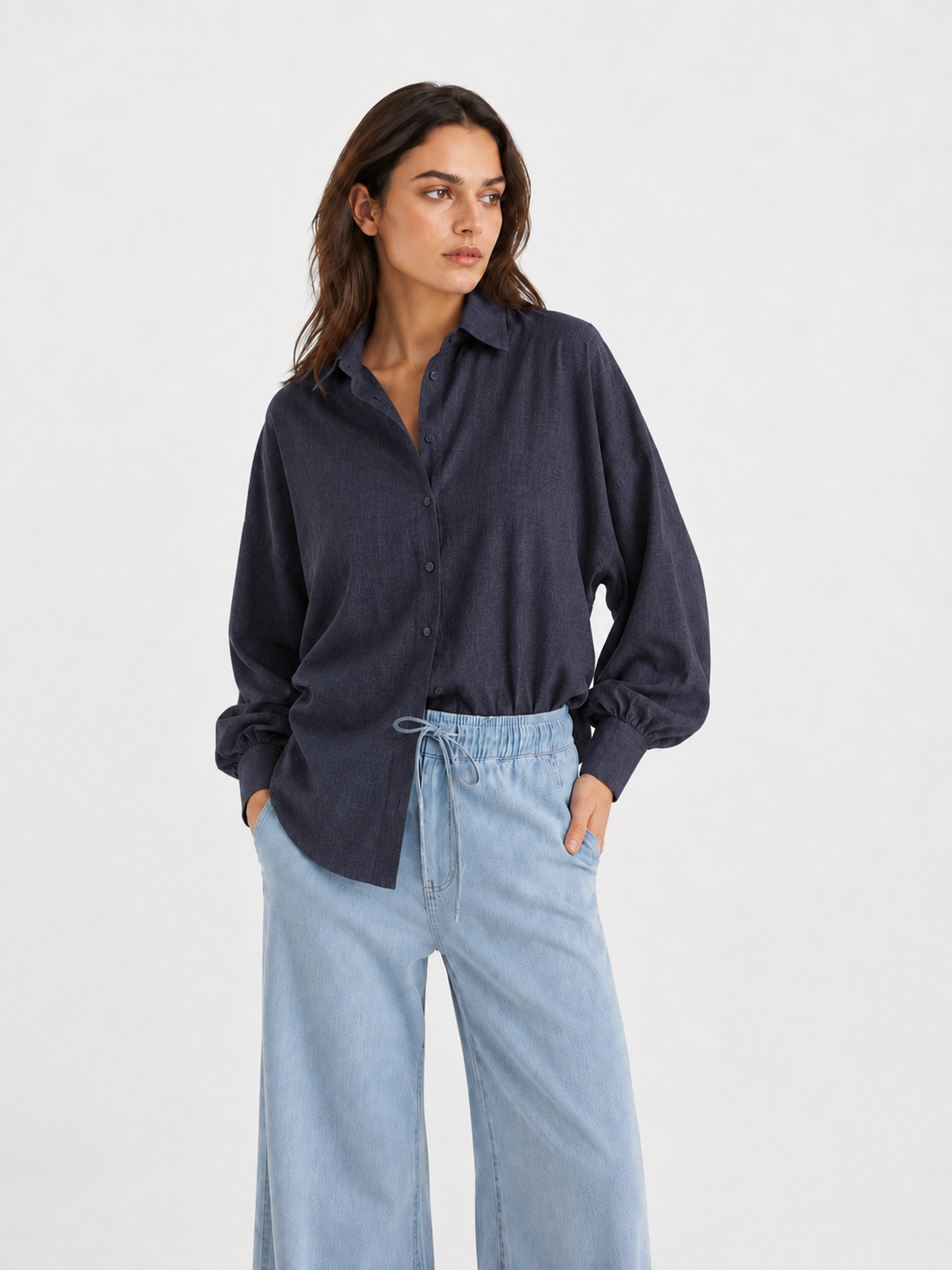Eine Frau, die ein eng anliegendes, dunkelanthrazitfarbenes Flanellhemd mit Knöpfen – ähnlich dem Modell „Bluse Antonia Slim Flannel Anthra“ – sowie eine hellblaue Hose mit weitem Bein trägt, steht mit den Händen in den Taschen vor einem schlichten, hellen Hintergrund und blickt zur Seite.