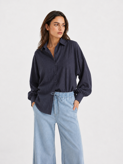 Eine Frau, die ein eng anliegendes, dunkelanthrazitfarbenes Flanellhemd mit Knöpfen – ähnlich dem Modell „Bluse Antonia Slim Flannel Anthra“ – sowie eine hellblaue Hose mit weitem Bein trägt, steht mit den Händen in den Taschen vor einem schlichten, hellen Hintergrund und blickt zur Seite.