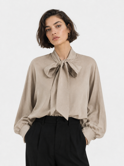 Eine Person mit kurzen braunen Haaren trägt die Bluse „Antonia Bow Taupe“, eine elegante Damenbluse in Taupe mit einer großen Schleife am Hals, dazu eine schwarze Hose, und steht vor einem schlichten weißen Hintergrund.
