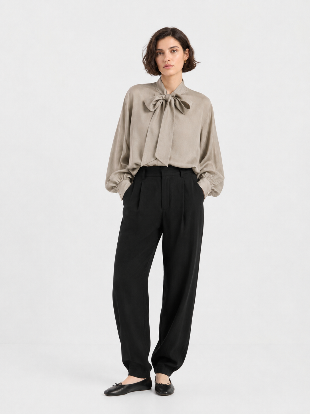 Vor einem schlichten Hintergrund steht eine Frau, die die „Bluse Antonia Bow Taupe“ trägt, eine elegante Damenbluse in Beige mit einer markanten großen Schleife am Hals – eine klassische Schleifenbluse –, kombiniert mit einer schwarzen Hose mit hoher Taille und schwarzen Ballerinas. Ihre Hände stecken in den Taschen, und sie hat kurzes, welliges braunes Haar.