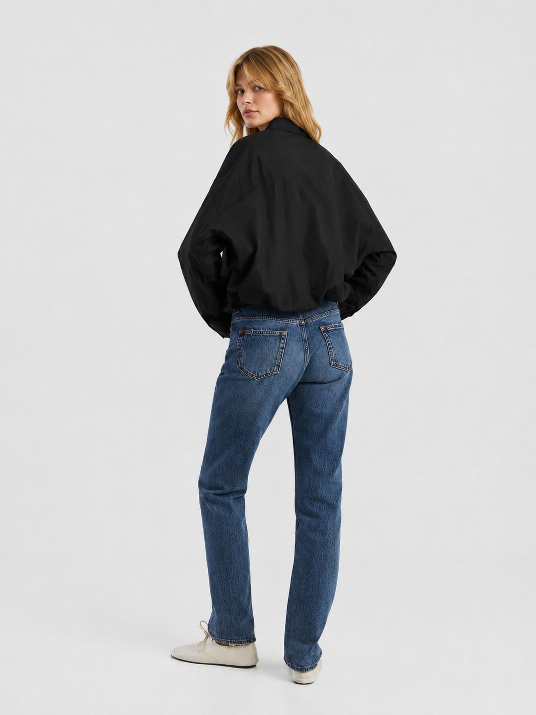 Eine Person mit blonden Haaren trägt eine schwarze Jacke, die „SIENNA Jeans Dark Blue – Straight Leg Damenjeans“ und weiße Schuhe; sie steht vor einem schlichten, hellen Hintergrund und blickt über die Schulter.