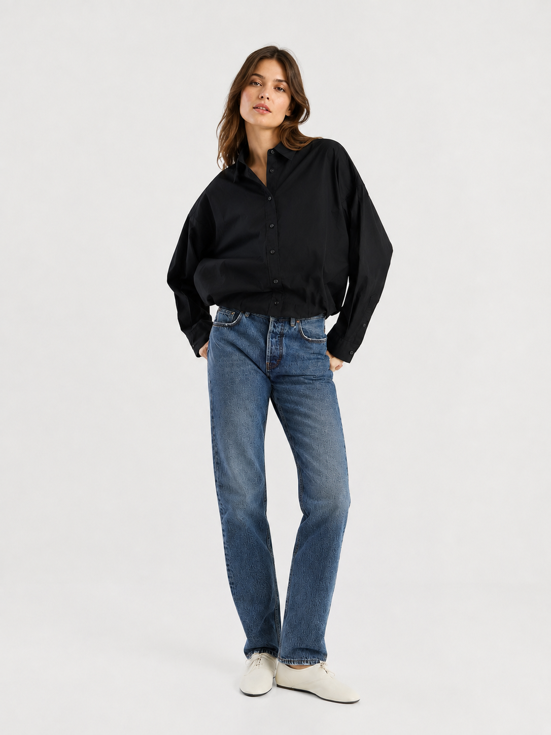 Eine Frau steht vor einem schlichten weißen Hintergrund, blickt leicht zur Seite und hat eine Hand in der Tasche. Sie trägt ein lockeres schwarzes Hemd mit Knöpfen, eine „SIENNA Jeans Dark Blue – Straight Leg Damenjeans“ und weiße Schuhe.