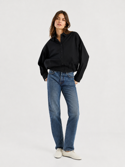 Eine Frau steht vor einem schlichten weißen Hintergrund, blickt leicht zur Seite und hat eine Hand in der Tasche. Sie trägt ein lockeres schwarzes Hemd mit Knöpfen, eine „SIENNA Jeans Dark Blue – Straight Leg Damenjeans“ und weiße Schuhe.