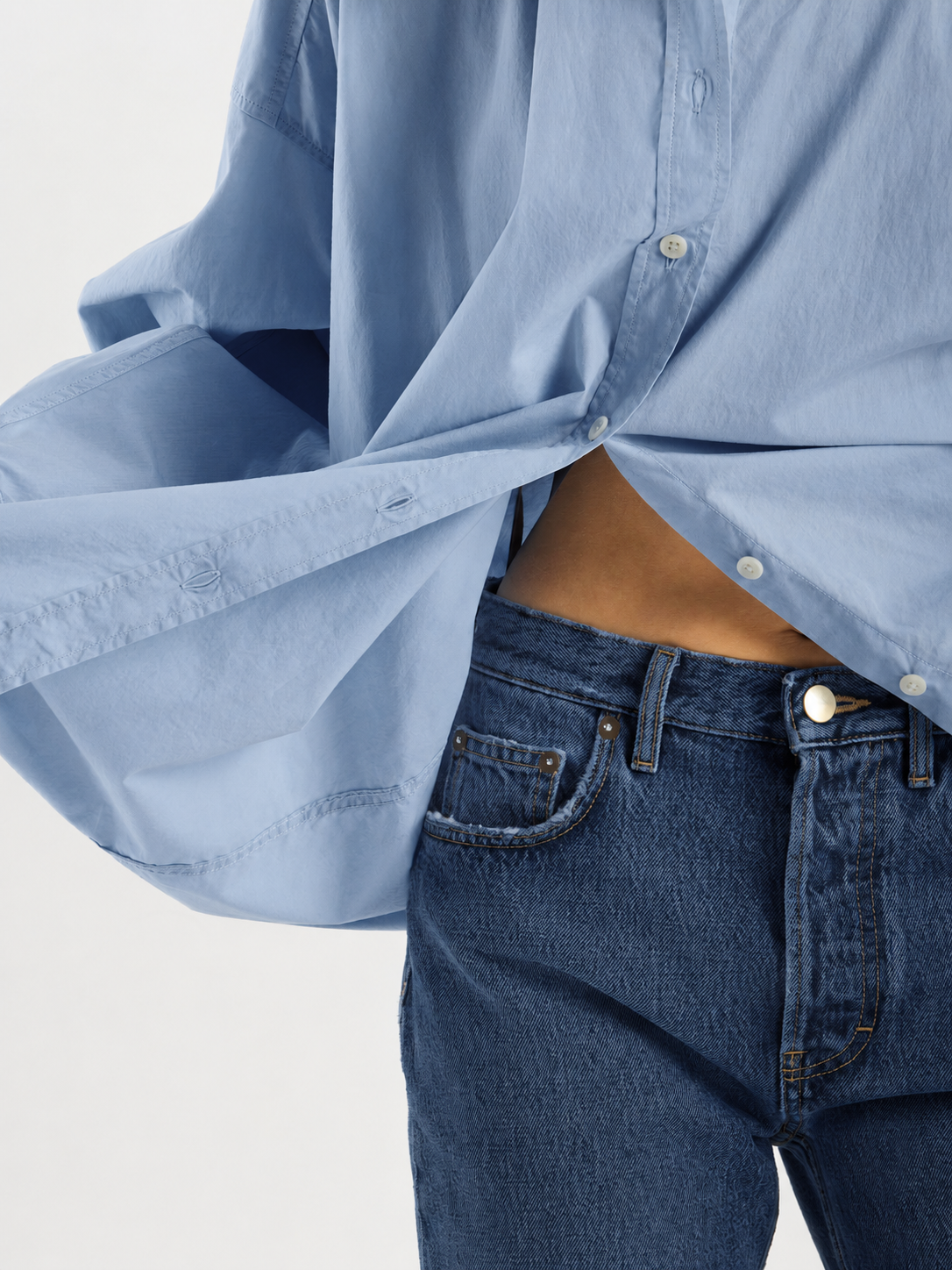 Eine Person, die eine dunkelblaue SIENNA-Jeans mit geradem Bein (Damenjeans) trägt, kombiniert mit einem hellblauen Hemd, das teilweise aufgeknöpft ist und nach außen weht. Das Foto ist so beschnitten, dass nur der Oberkörper zu sehen ist.