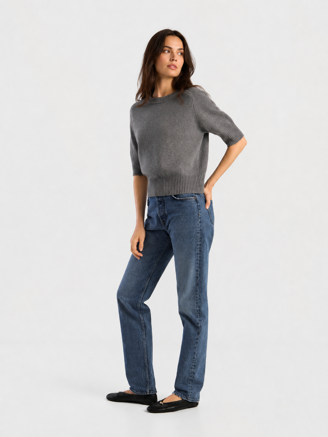 Eine Person, die einen grauen Kurzarmpullover, eine NOLA Jeans Light Blue – Wide Leg Damenjeans und schwarze Ballerinas trägt, steht mit einer Hand in der Tasche und blickt vor einem schlichten hellen Hintergrund zur Seite.