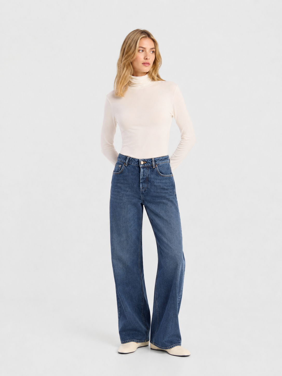 Eine Frau steht vor einem hellgrauen Hintergrund, blickt zur Seite, die Hände hinter dem Rücken verschränkt, und trägt einen weißen Langarm-Rollkragenpullover, eine hellblaue NOLA-Jeans mit weitem Bein (Damenjeans) sowie weiße Schuhe.