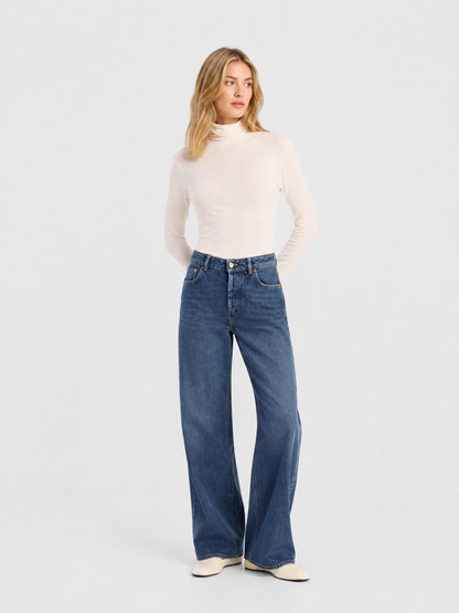 Eine Frau steht vor einem hellgrauen Hintergrund, blickt zur Seite, die Hände hinter dem Rücken verschränkt, und trägt einen weißen Langarm-Rollkragenpullover, eine hellblaue NOLA-Jeans mit weitem Bein (Damenjeans) sowie weiße Schuhe.